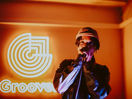 Eine Person singt in ein Mikrofon vor einem leuchtenden Groove-Logo an einer orange beleuchteten Wand.