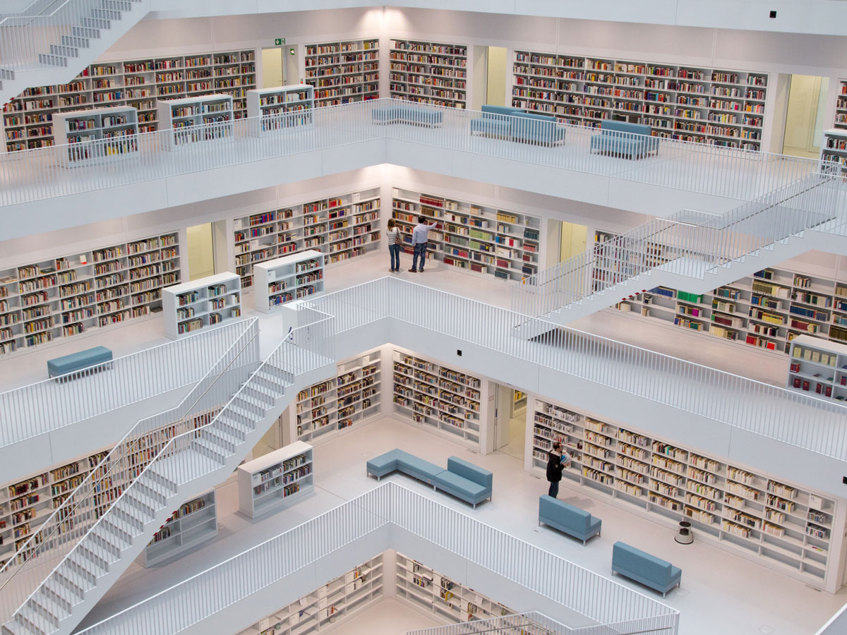 Moderne Bibliothek mit weißen Regalen, Treppen und Sitzbänken, gefüllt mit Büchern, Menschen stöbern.