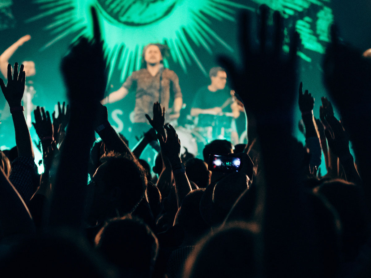 Menschenmenge bei Konzert, hebt Hände vor beleuchteter Bühne mit Musikern.