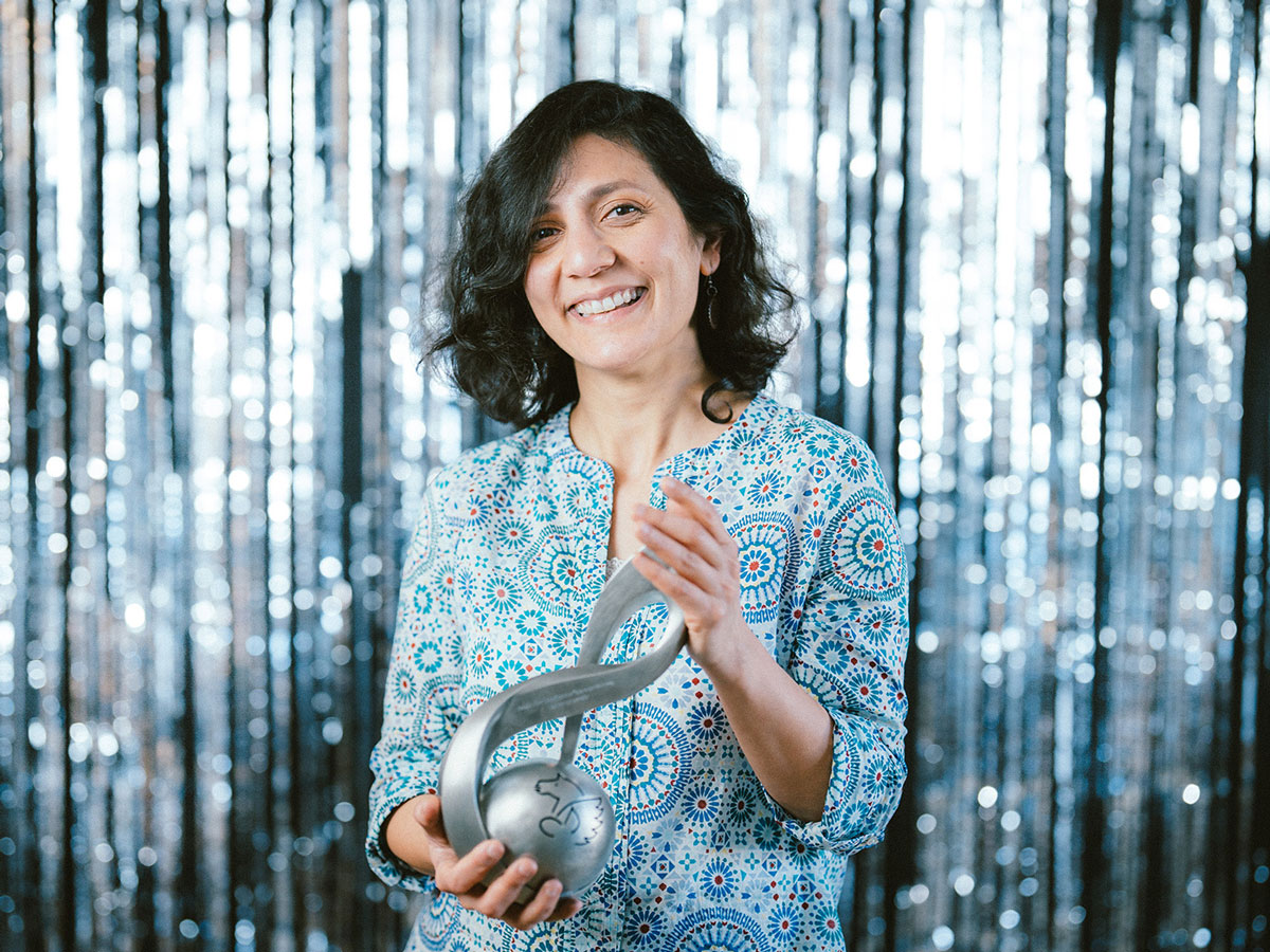 Eine lächelnde Person hält eine silberne Trophäe vor einem glitzernden Hintergrund.