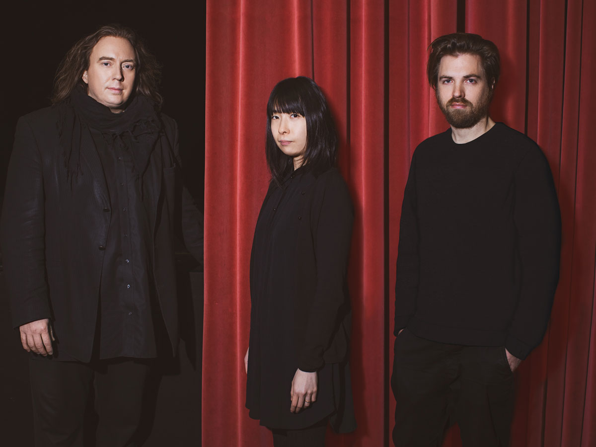 Drei Personen in schwarzer Kleidung vor einem roten Vorhang, eine links im Dunkeln, zwei rechts vor dem Vorhang.