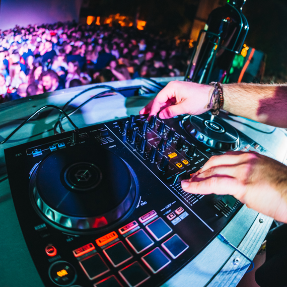 DJ mixt Musik an einem Mischpult, während eine große Menschenmenge im Hintergrund tanzt.