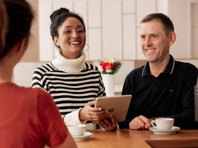 Drei Personen sitzen lächelnd an einem Tisch mit Tassen, eine Frau hält ein Tablet.