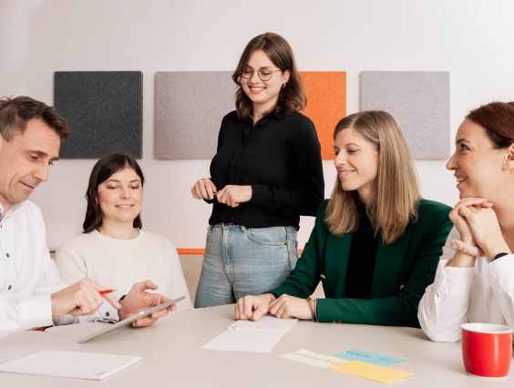 Fünf Personen in einem Meeting, lächelnd, mit Notizen und Tablet an einem Tisch.