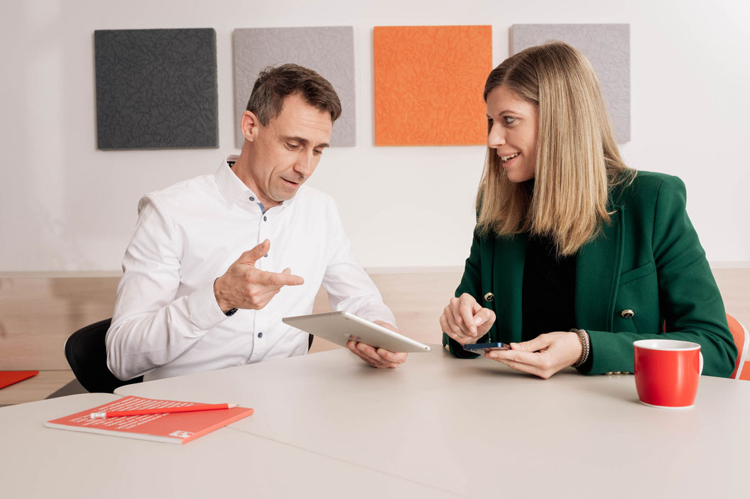 Ein Mann und eine Frau diskutieren an einem Tisch mit Tablet und Smartphone, rote Tasse und Notizbuch.