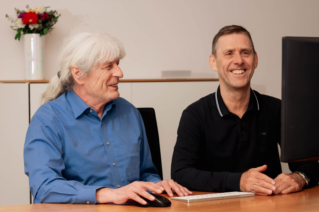 Zwei Männer sitzen lächelnd vor einem Computer, im Hintergrund steht eine Vase mit bunten Blumen.