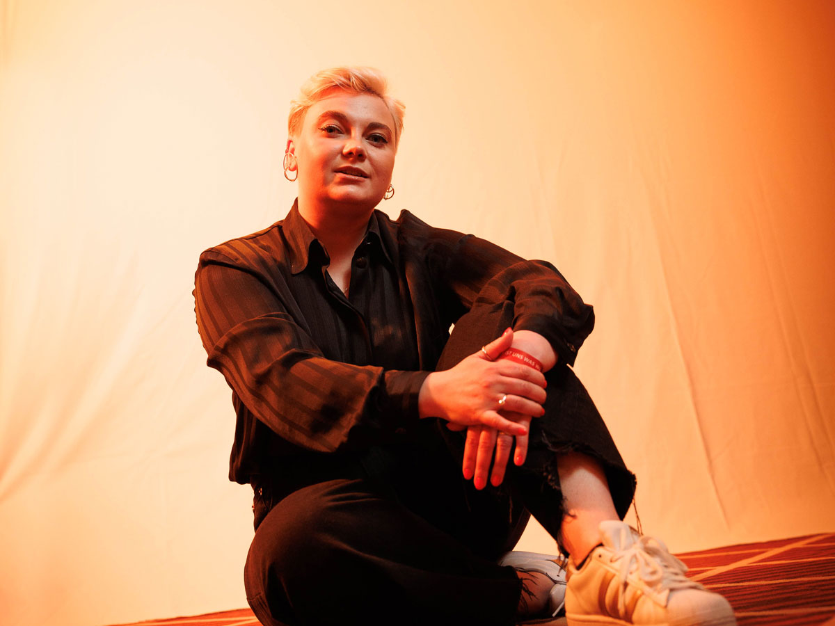 Person in schwarzem Outfit sitzt auf gestreiftem Boden vor einem orange beleuchteten Hintergrund.