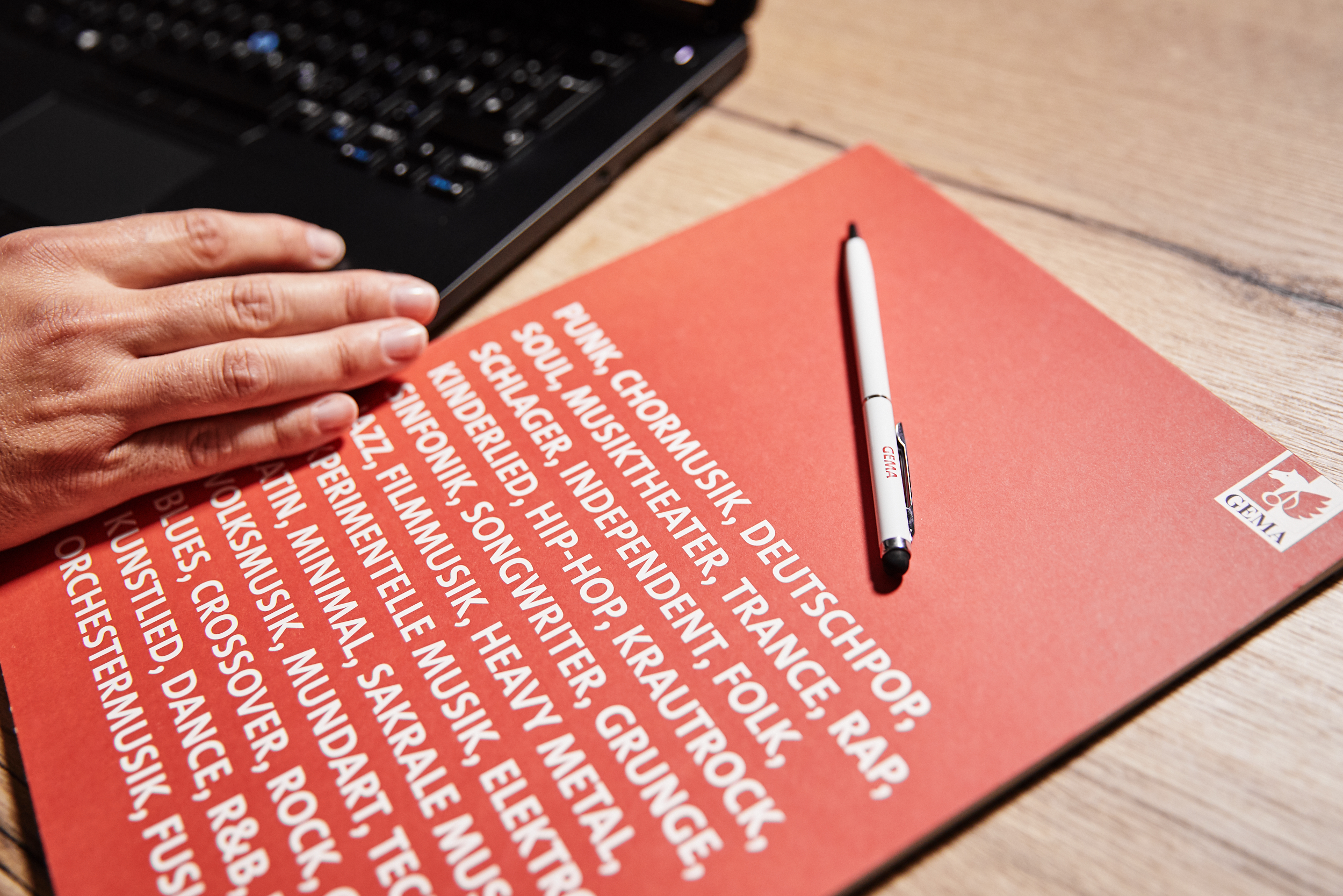 Eine Hand auf einer Laptop-Tastatur neben einem roten Papier mit Musikgenres und einem weißen Stift.