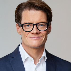 Ein Mann mit Brille in Anzug lächelt vor neutralem Hintergrund.