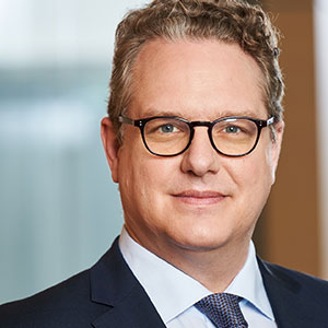 Eine Person im Anzug und mit Brille lächelt leicht und steht in einem Innenraum vor einem unscharfen Hintergrund.