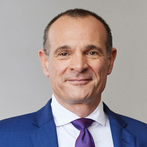 Ein Mann im blauen Anzug mit lila Krawatte vor grauem Hintergrund.