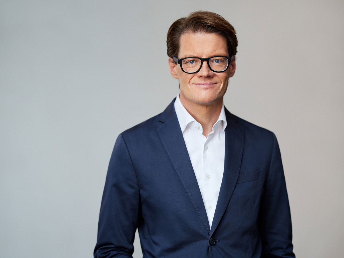 Ein Mann in Anzug und Brille lächelt vor neutralem Hintergrund.