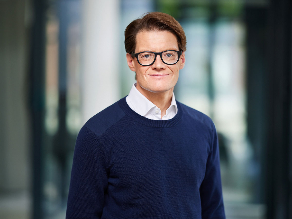 Ein Mann mit Brille und blauem Pullover steht vor einem unscharfen Hintergrund.