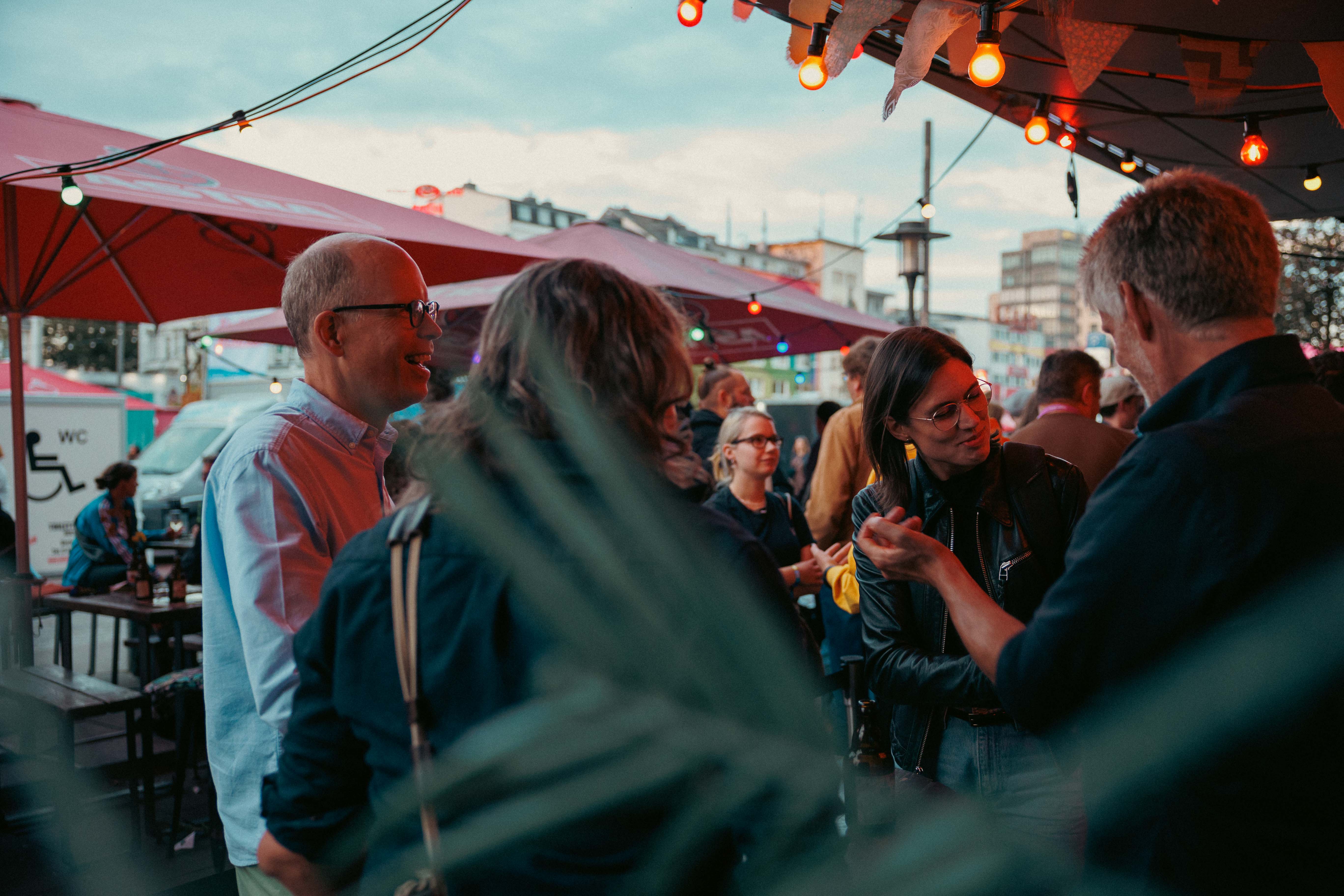 Eine Gruppe von Menschen unterhält sich fröhlich auf einer Terrasse mit roten Sonnenschirmen und Lichterketten.