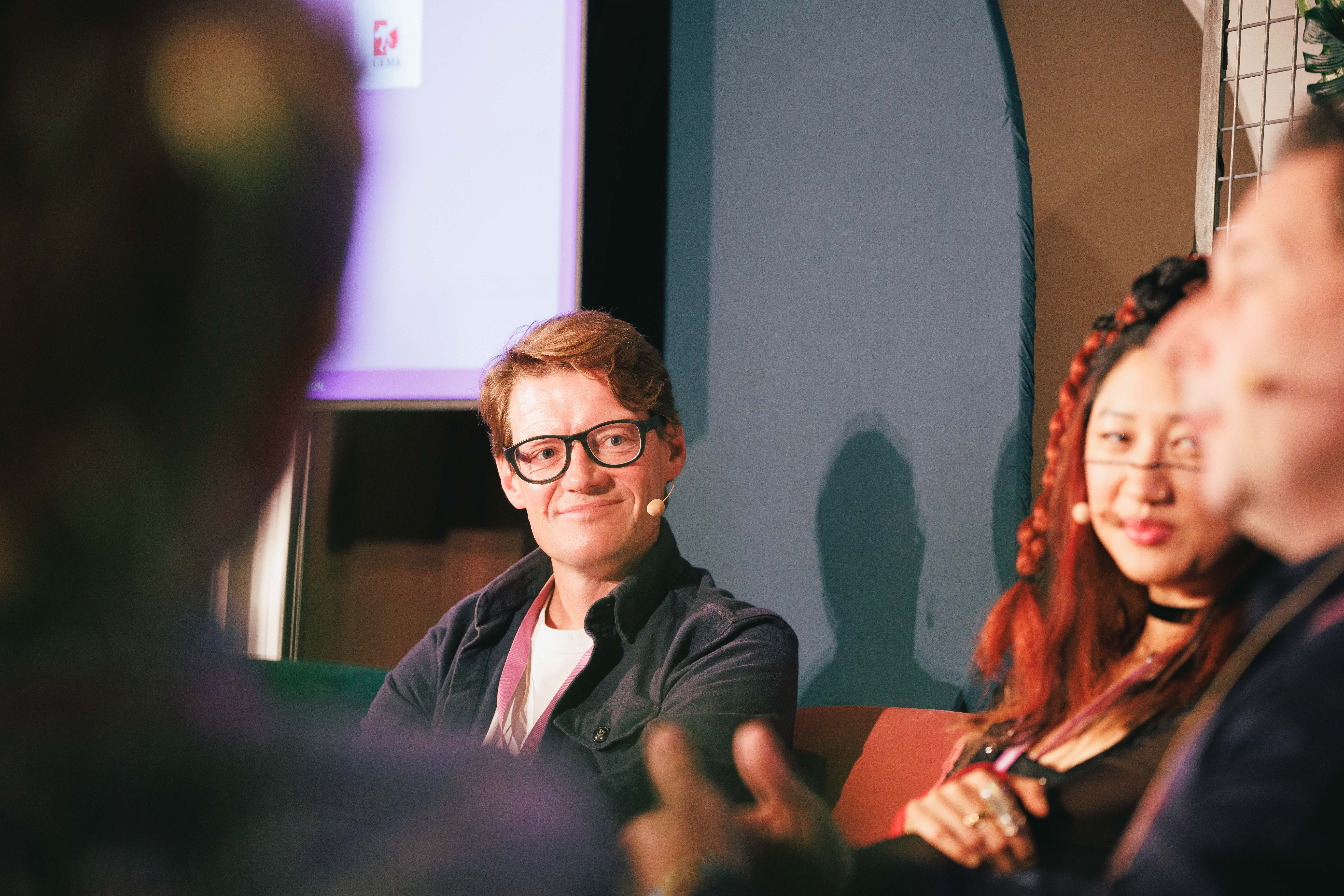 Eine Person mit Brille lächelt auf einem Podium, neben einer weiteren Person mit rotem Haar.