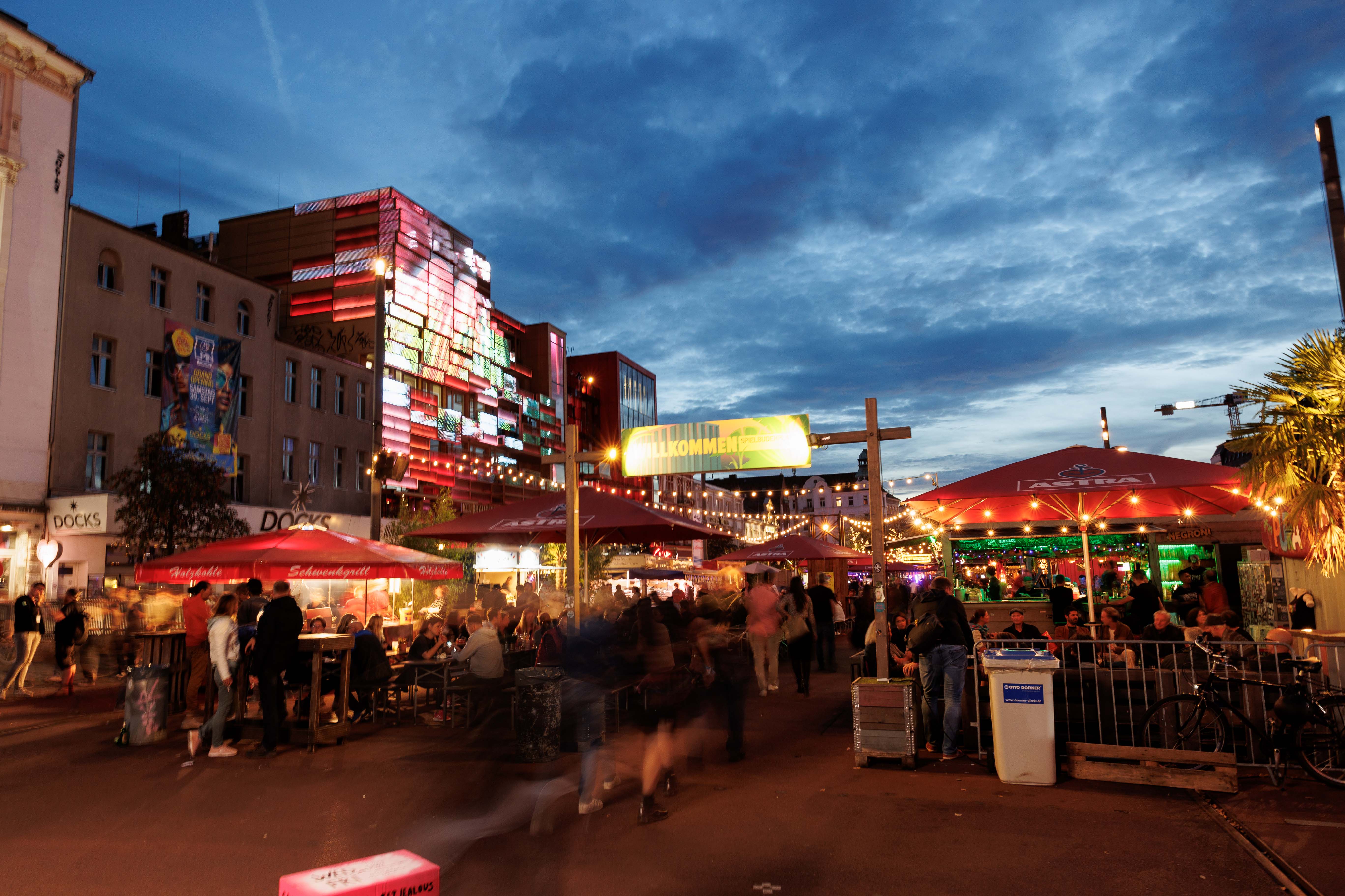 Nachtmarkt mit bunten Lichtern, Menschen unter roten Schirmen, beleuchtetes Gebäude im Hintergrund, blauer Himmel.