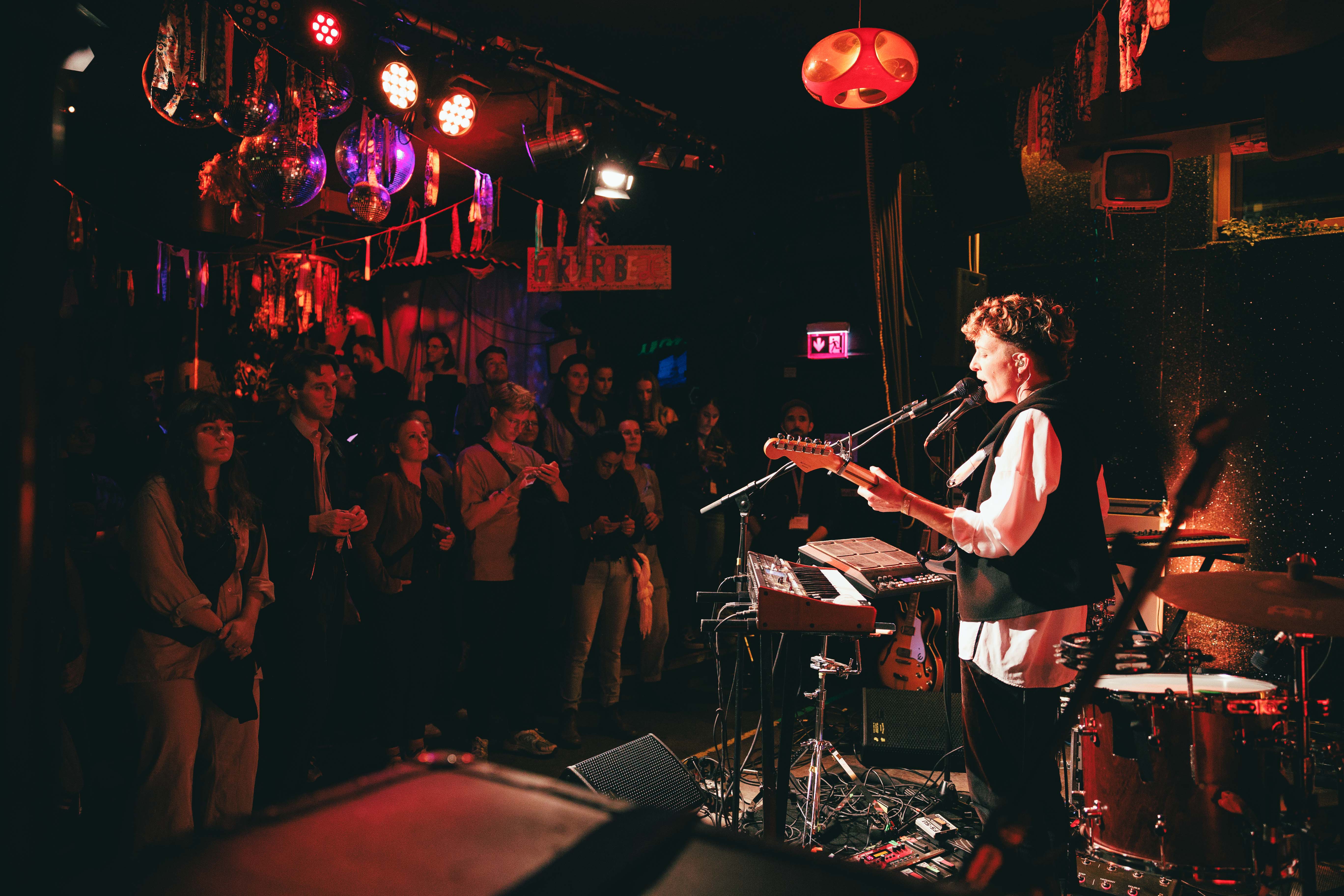 Ein Musiker spielt Gitarre auf einer Bühne, umgeben von rotem Licht, vor einem aufmerksamen Publikum in einem Club.