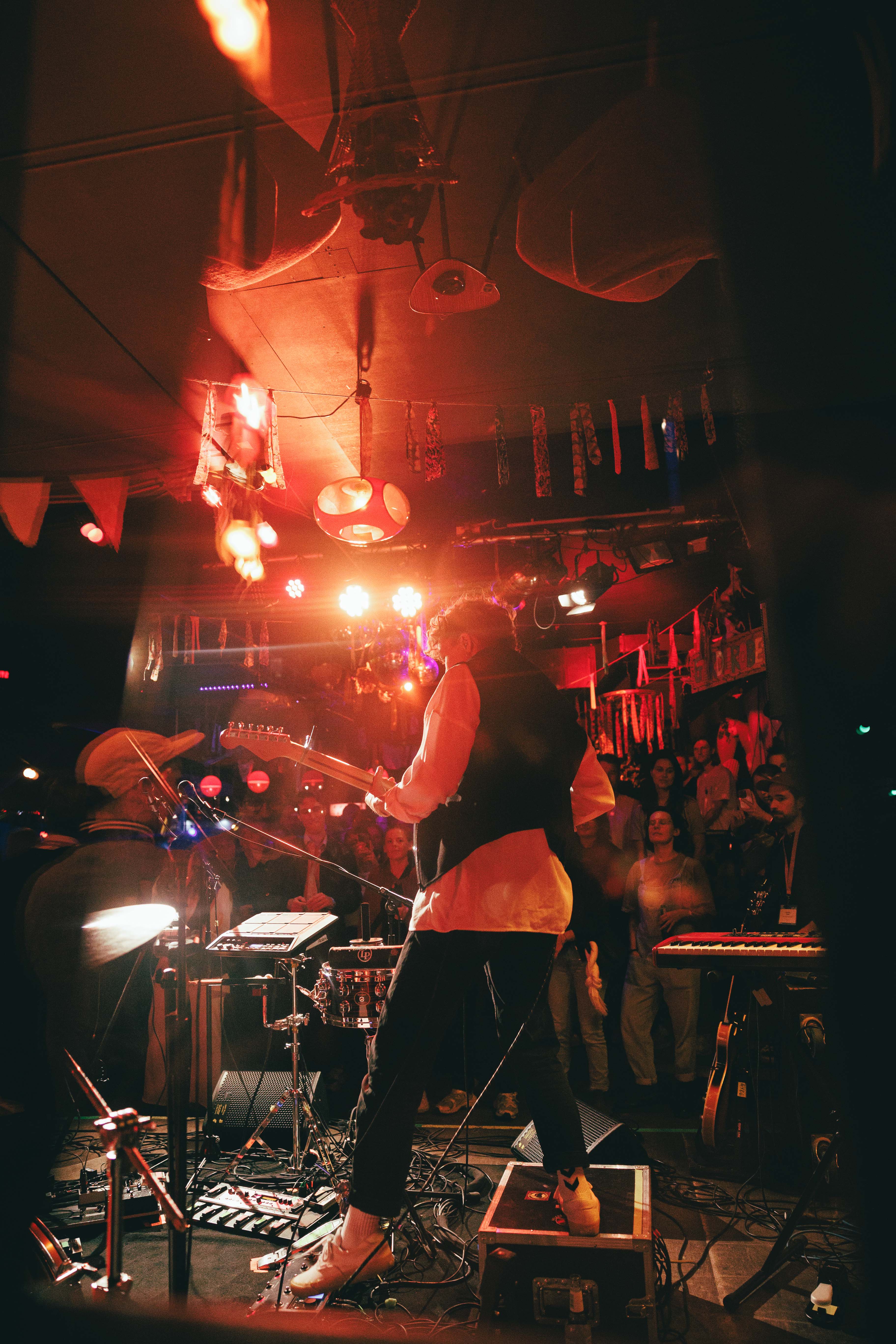 Eine Band spielt auf einer Bühne in einem Club mit roter Beleuchtung und Publikum im Hintergrund.