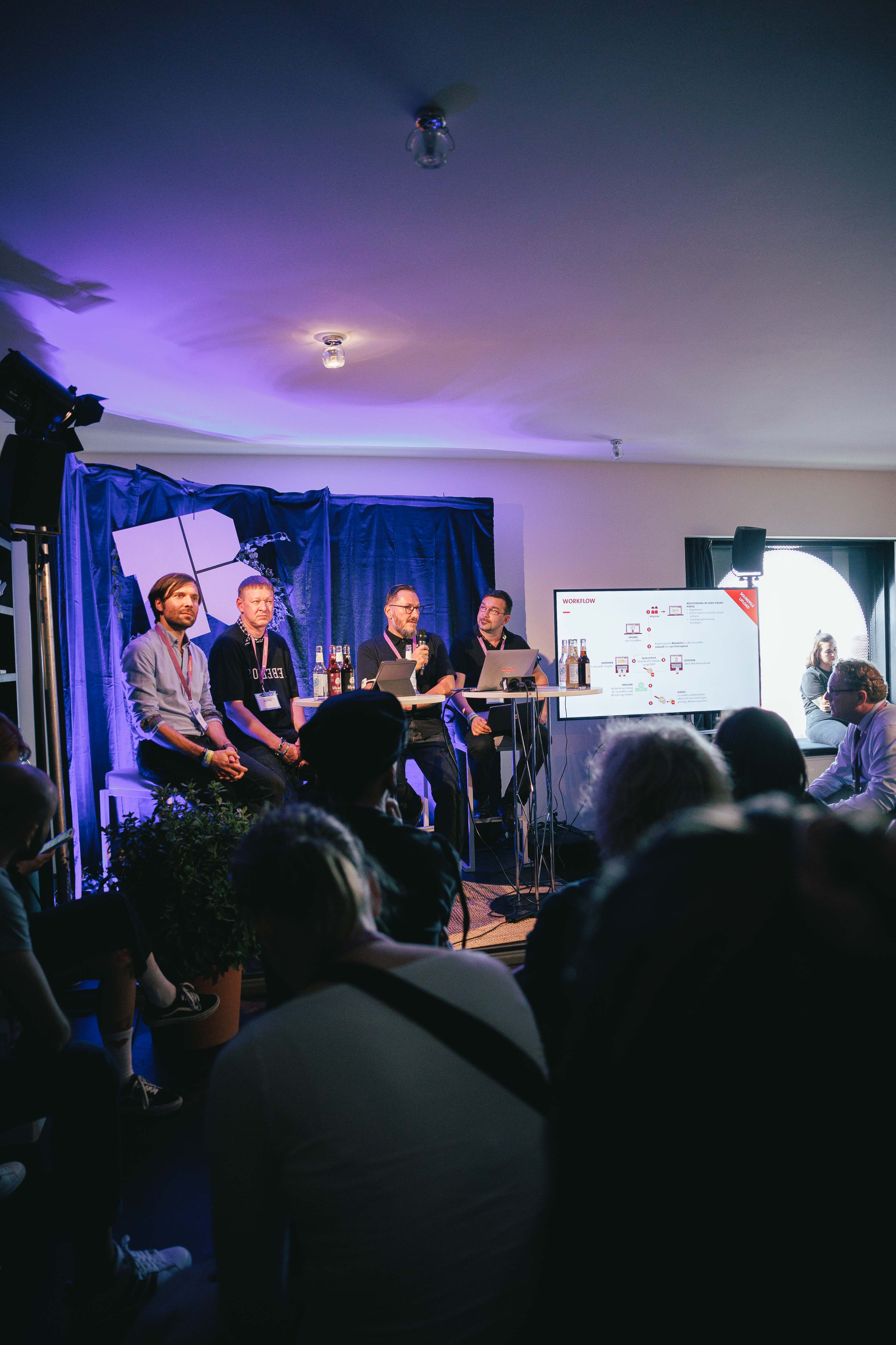 Vier Männer sitzen auf einer Bühne mit blauer Beleuchtung, Publikum im Vordergrund. Ein Bildschirm zeigt eine Präsentation.