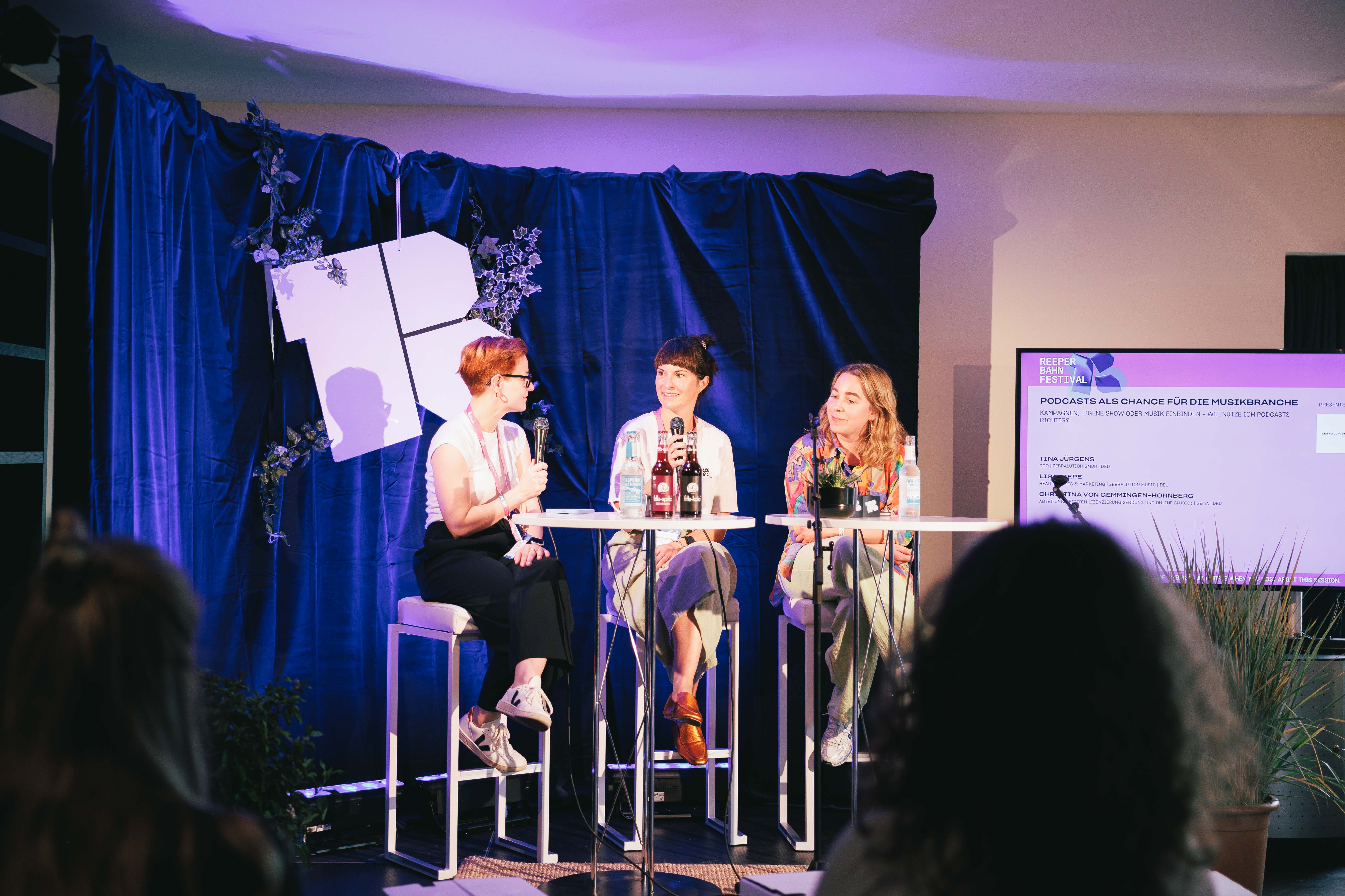 Drei Personen diskutieren auf einer Bühne mit blauer Kulisse, Mikrofone und Getränke auf einem Tisch.