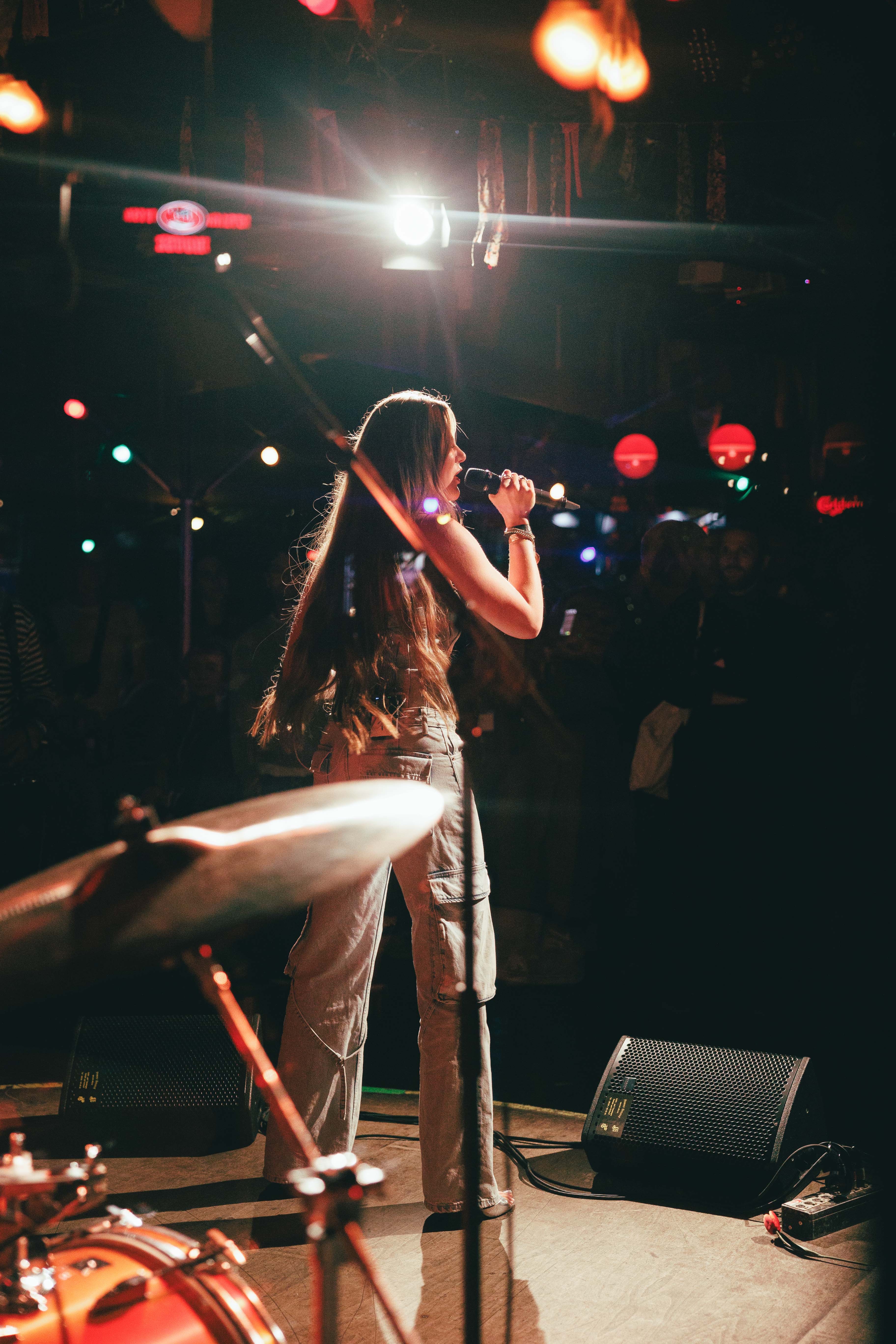Eine Frau singt auf einer Bühne, beleuchtet von bunten Lichtern, mit einem Schlagzeug im Vordergrund.