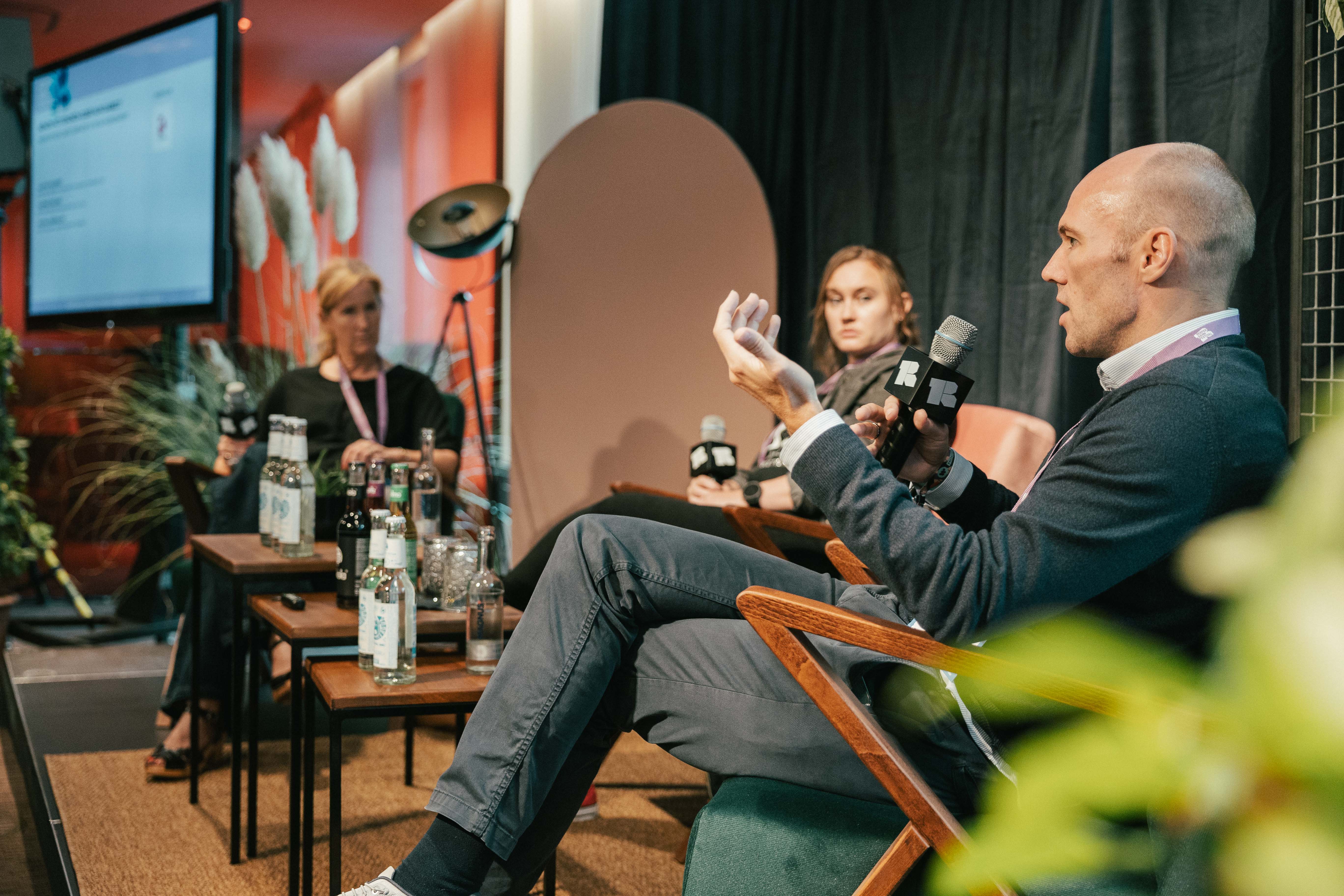 Drei Personen diskutieren auf einer Bühne, Mikrofone in der Hand, mit Getränken auf einem Tisch.