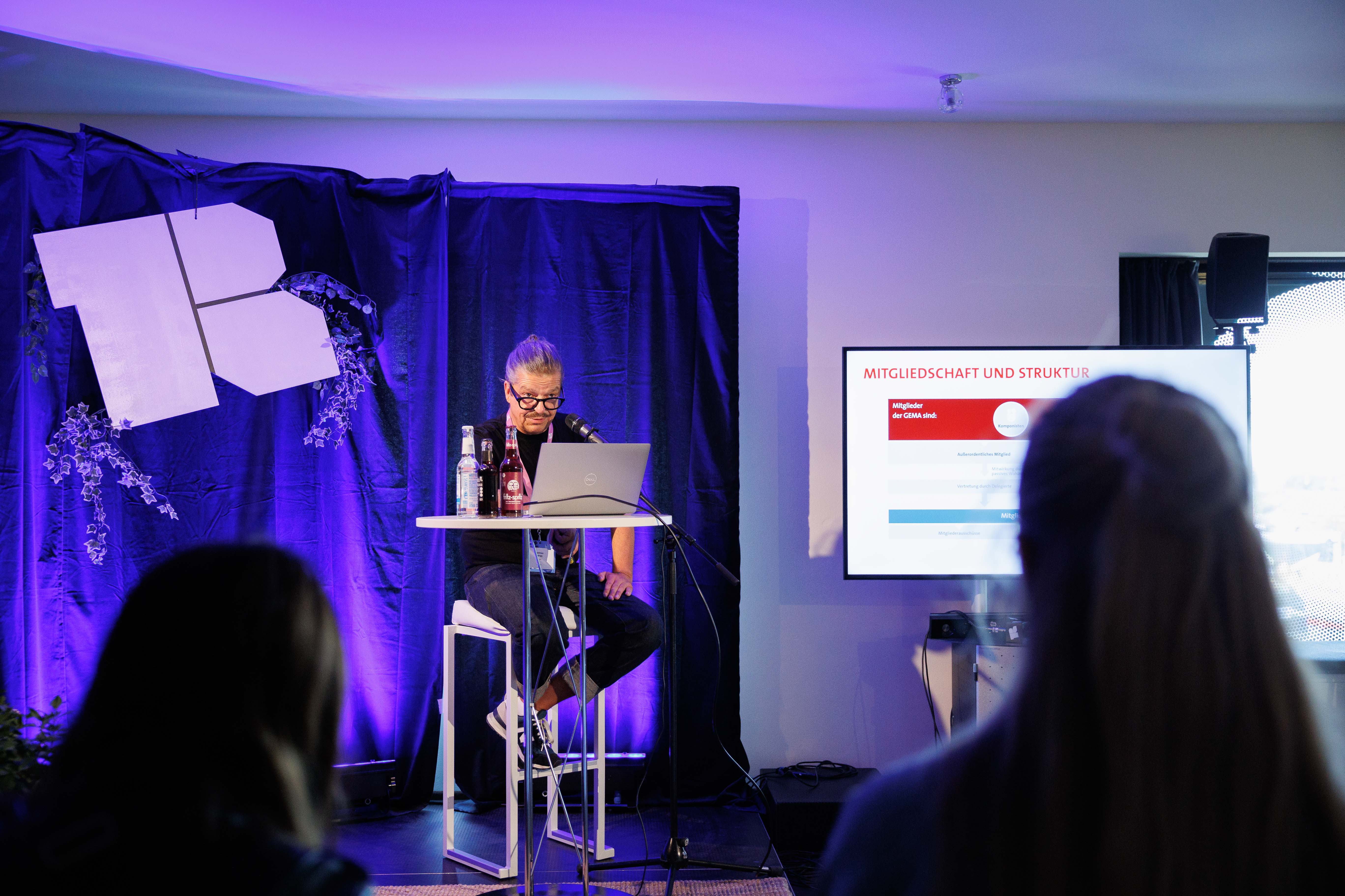 Eine Person sitzt auf einer Bühne vor einem Laptop, mit einem Bildschirm rechts und violetter Beleuchtung.