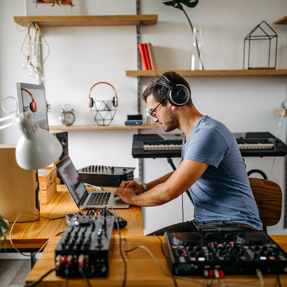 Ein Mann mit Kopfhörern arbeitet an einem Laptop, umgeben von Musikgeräten in einem modernen Raum.