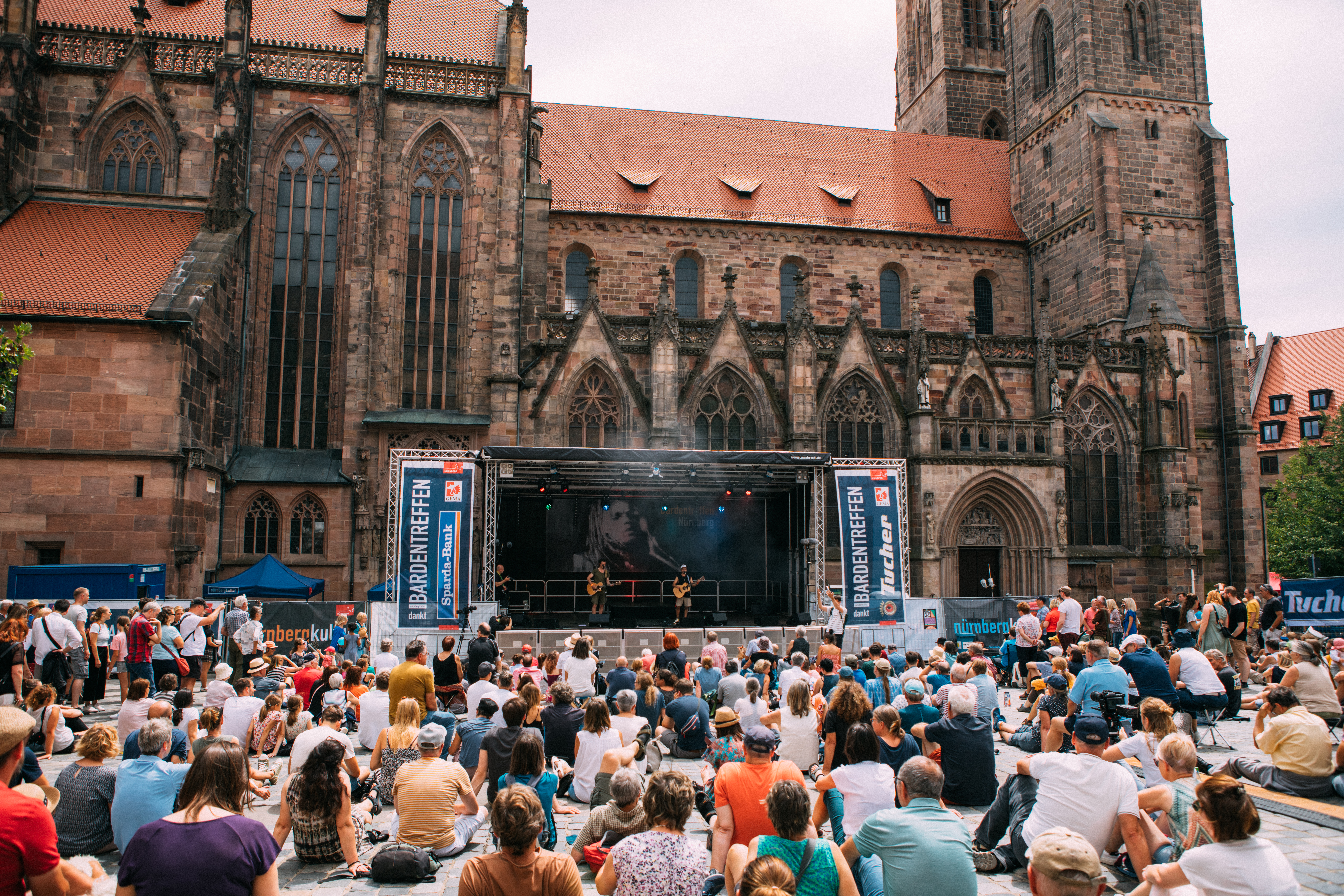 Menschenmenge sitzt vor einer Bühne mit Musikern, vor einer gotischen Kirche.