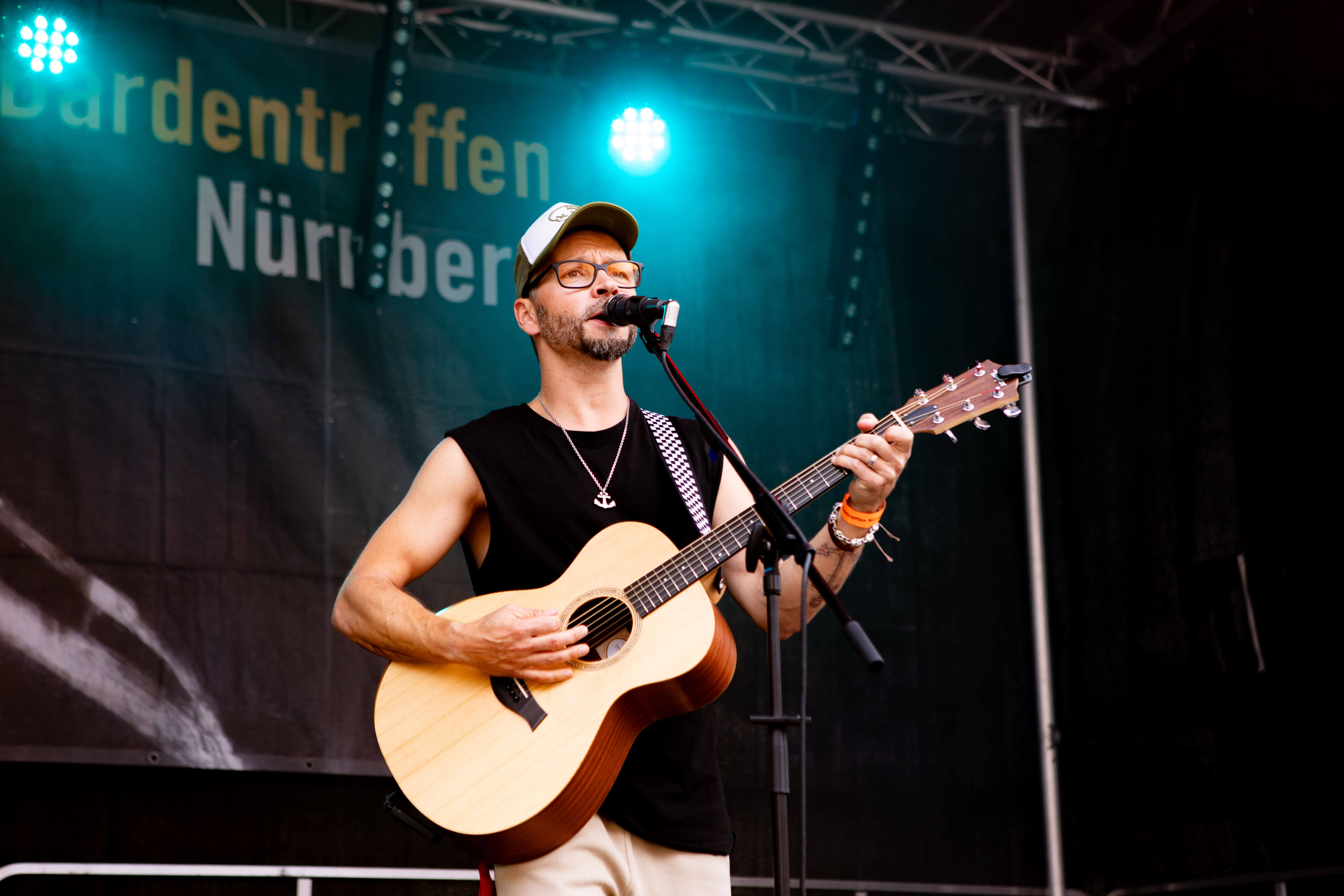 Mann mit Gitarre singt auf Bühne, trägt Kappe und Brille, Hintergrund mit Schriftzug 