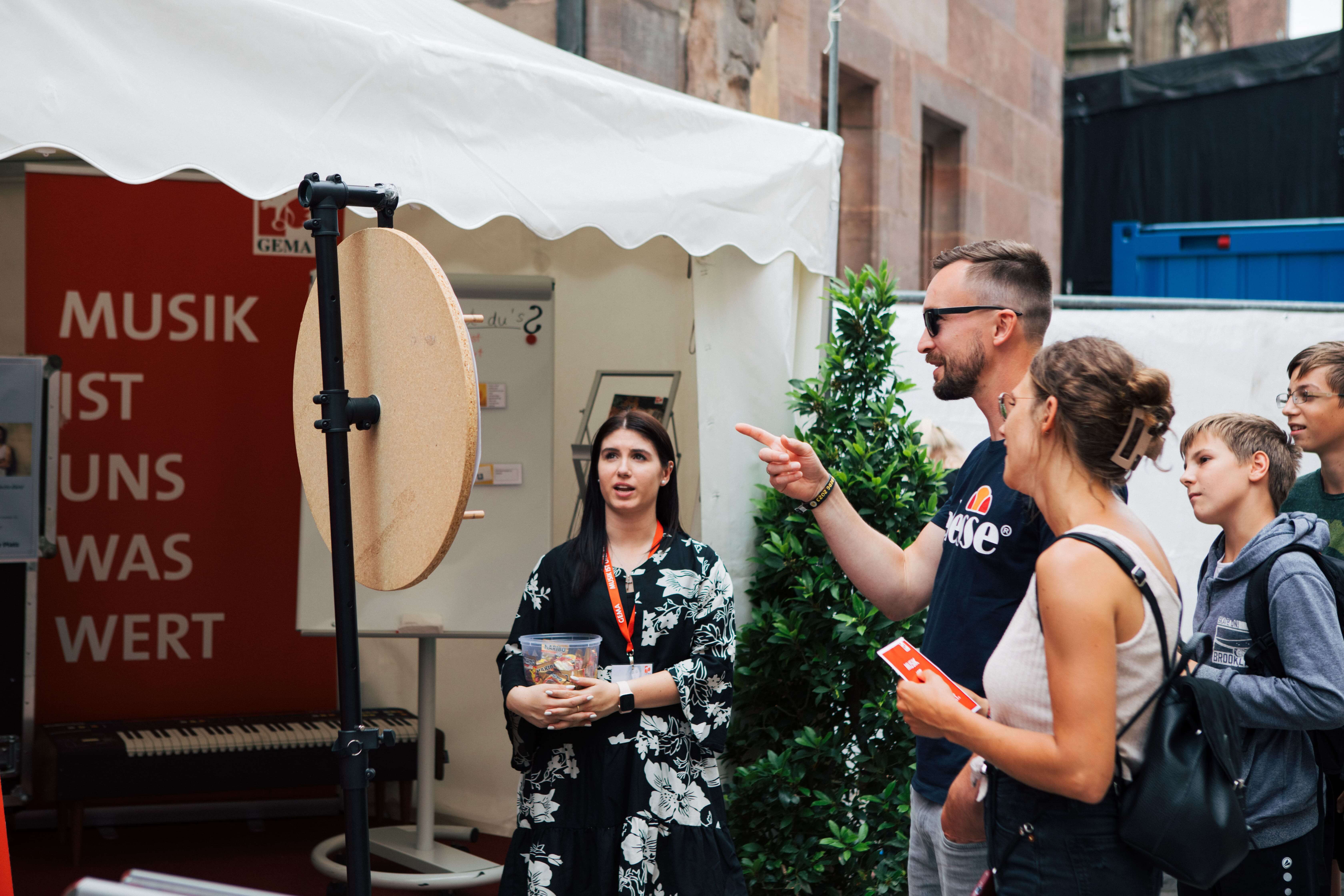 Gruppe von Menschen bei einem Glücksradspiel vor einem Zelt mit Pflanzen und rotem Plakat im Hintergrund.