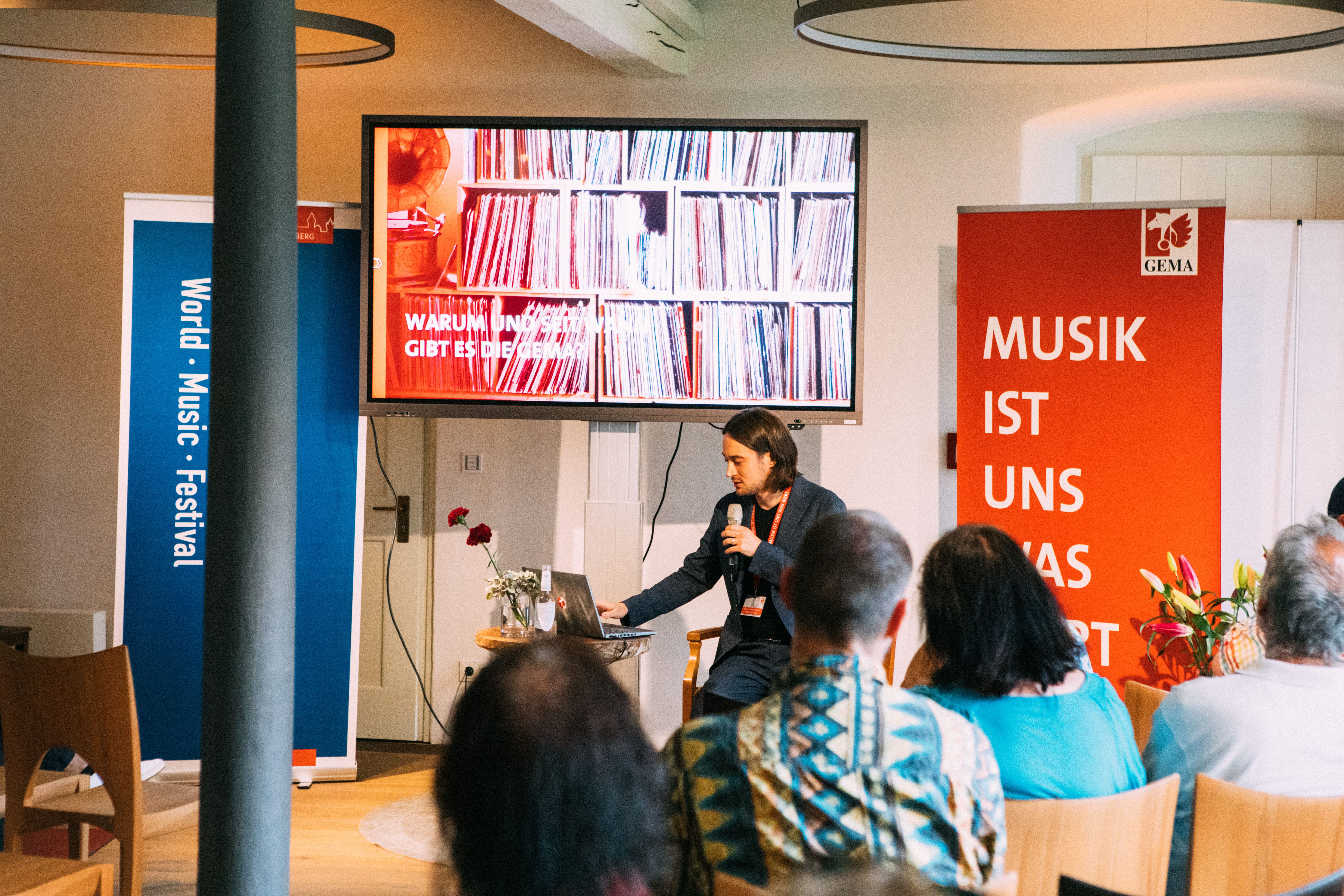 Ein Mann hält ein Mikrofon vor einem Bildschirm mit Plattenregalen, umgeben von Festivalbannern und Publikum.