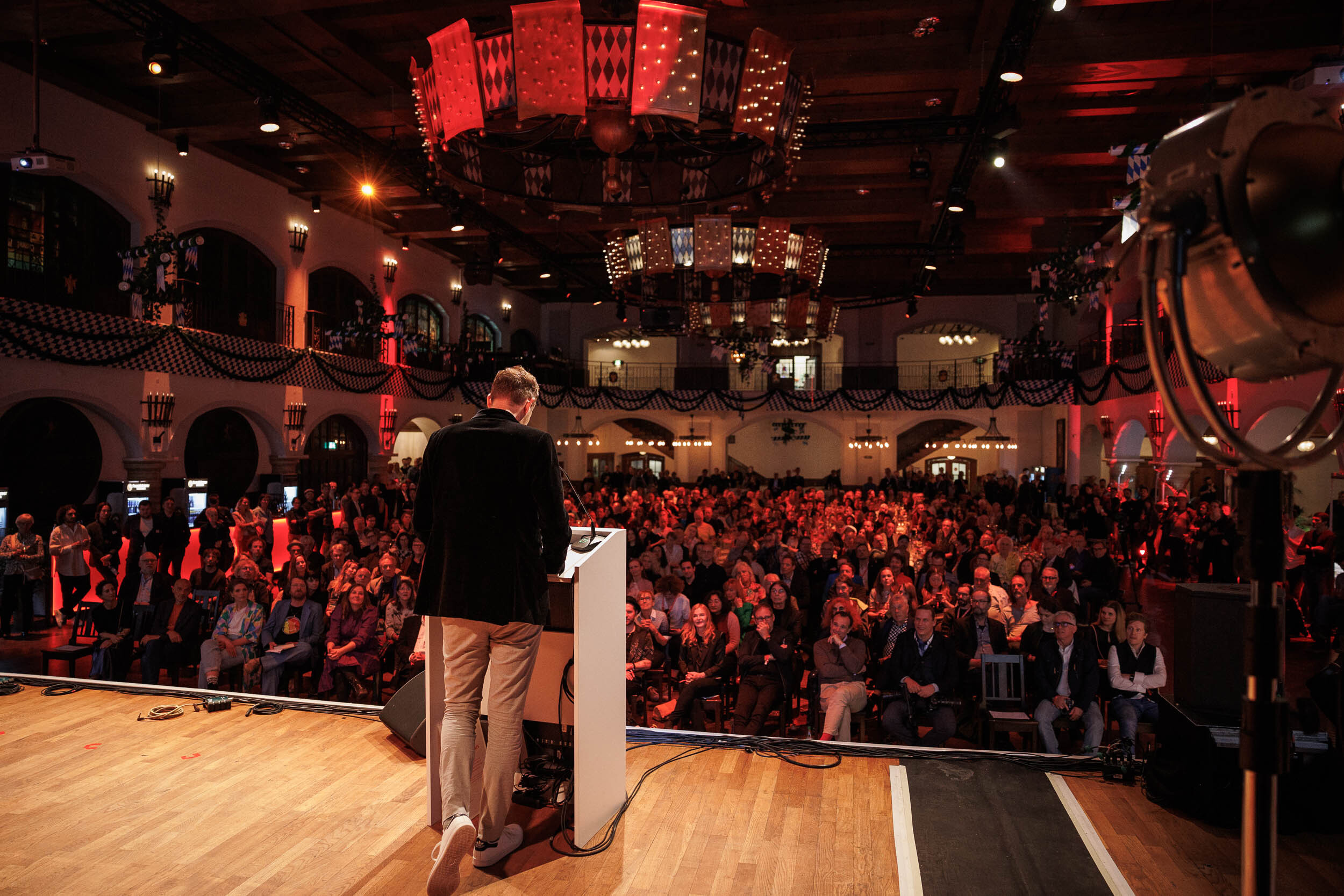 Eine Person spricht auf einer Bühne vor einem großen, aufmerksamen Publikum in einem festlich beleuchteten Saal.