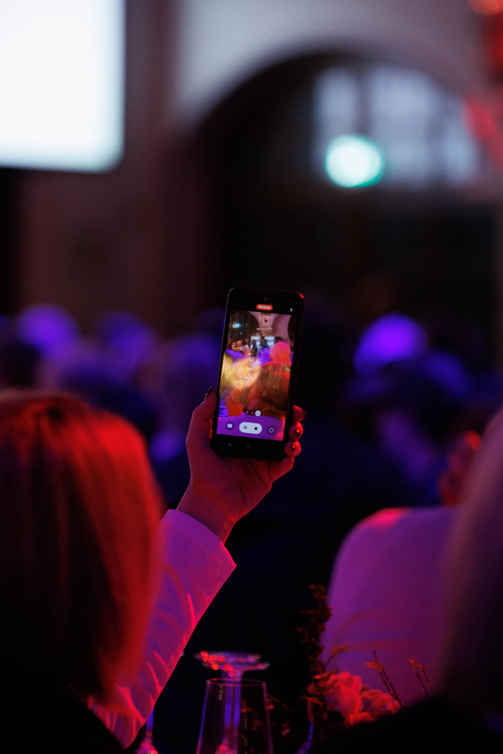 Eine Hand hält ein Smartphone, das ein buntes Event in einem dunklen Raum filmt.