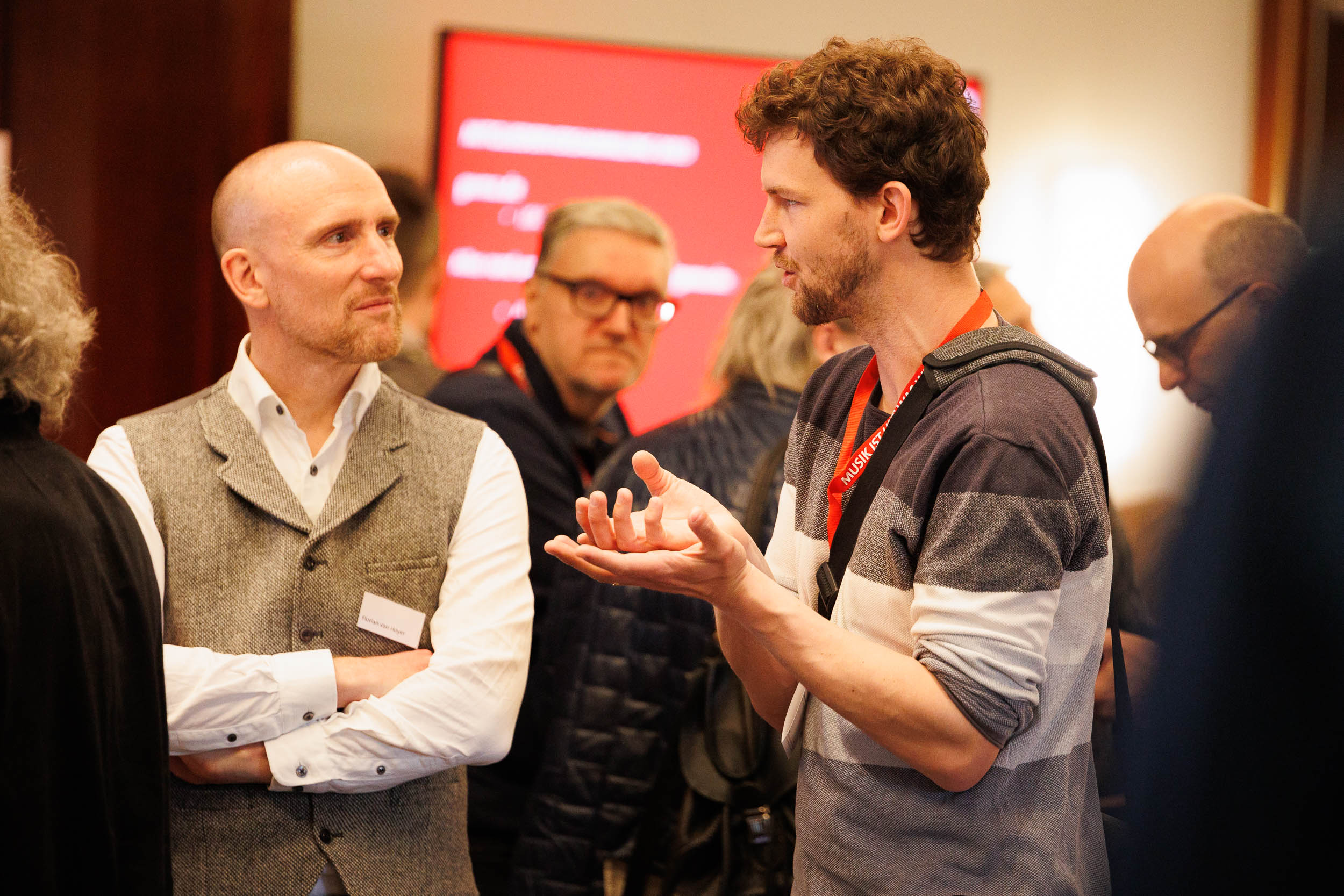 Zwei Männer im Gespräch, umgeben von anderen Personen, vor einem roten Bildschirm im Hintergrund.