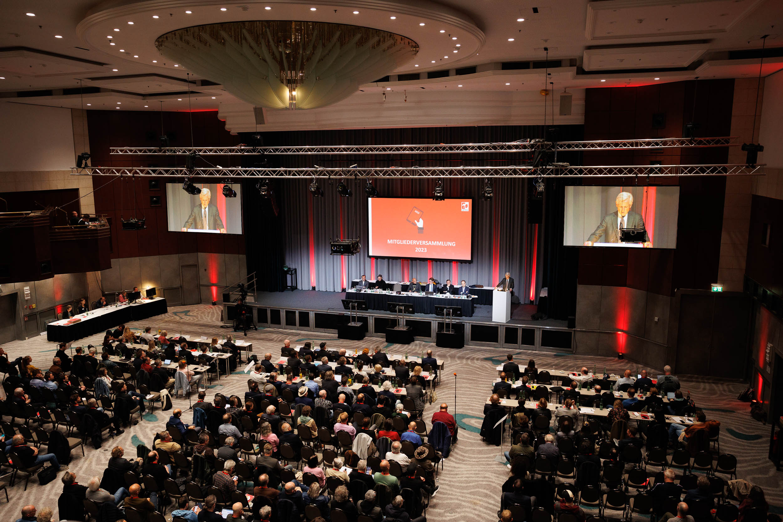Großer Konferenzsaal mit Publikum, Bühne, Rednerpult und Leinwand, die eine Versammlung zeigt.