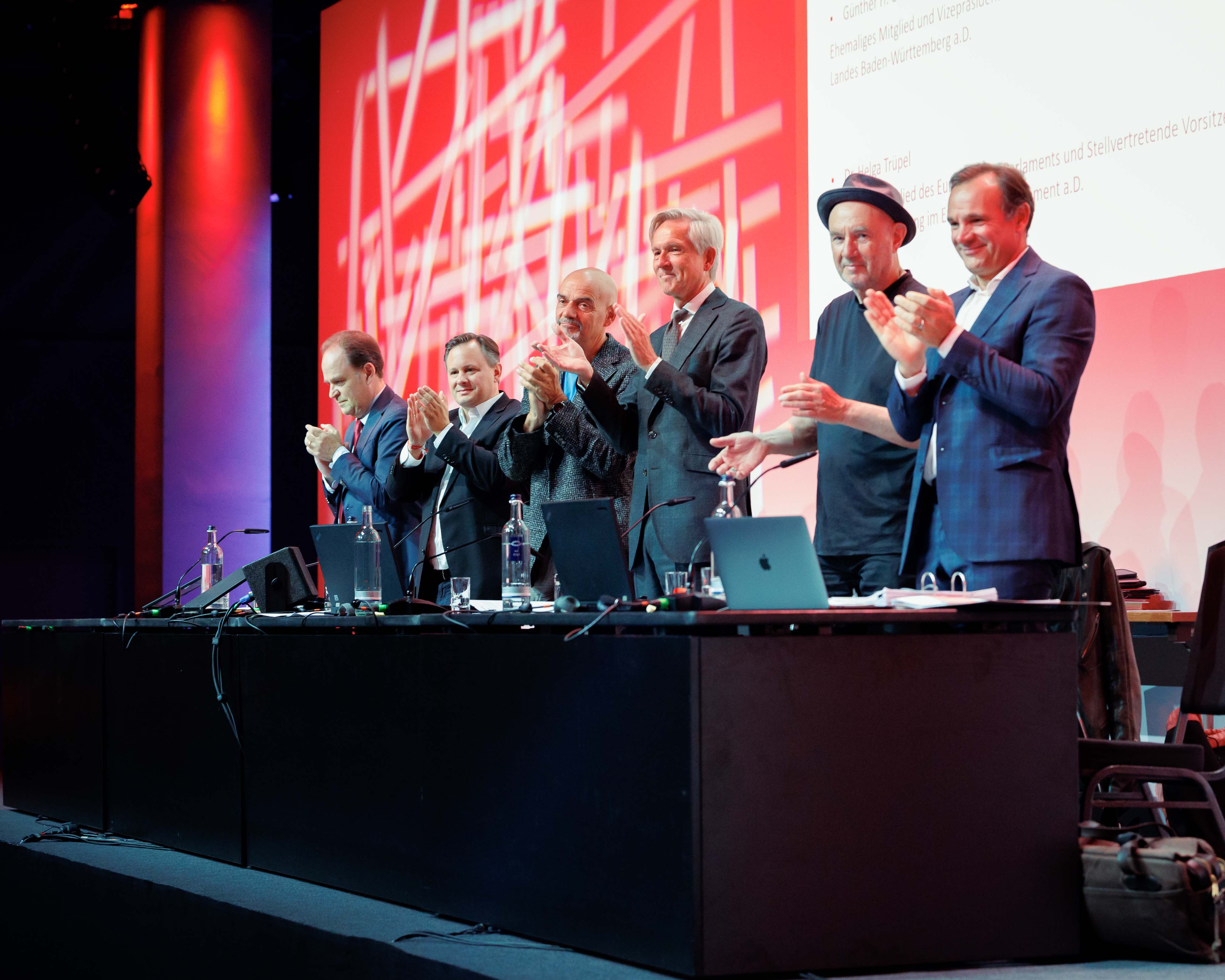 Sechs Männer in Anzügen und Hemden stehen klatschend hinter einem Tisch mit Laptops vor rotem Hintergrund.