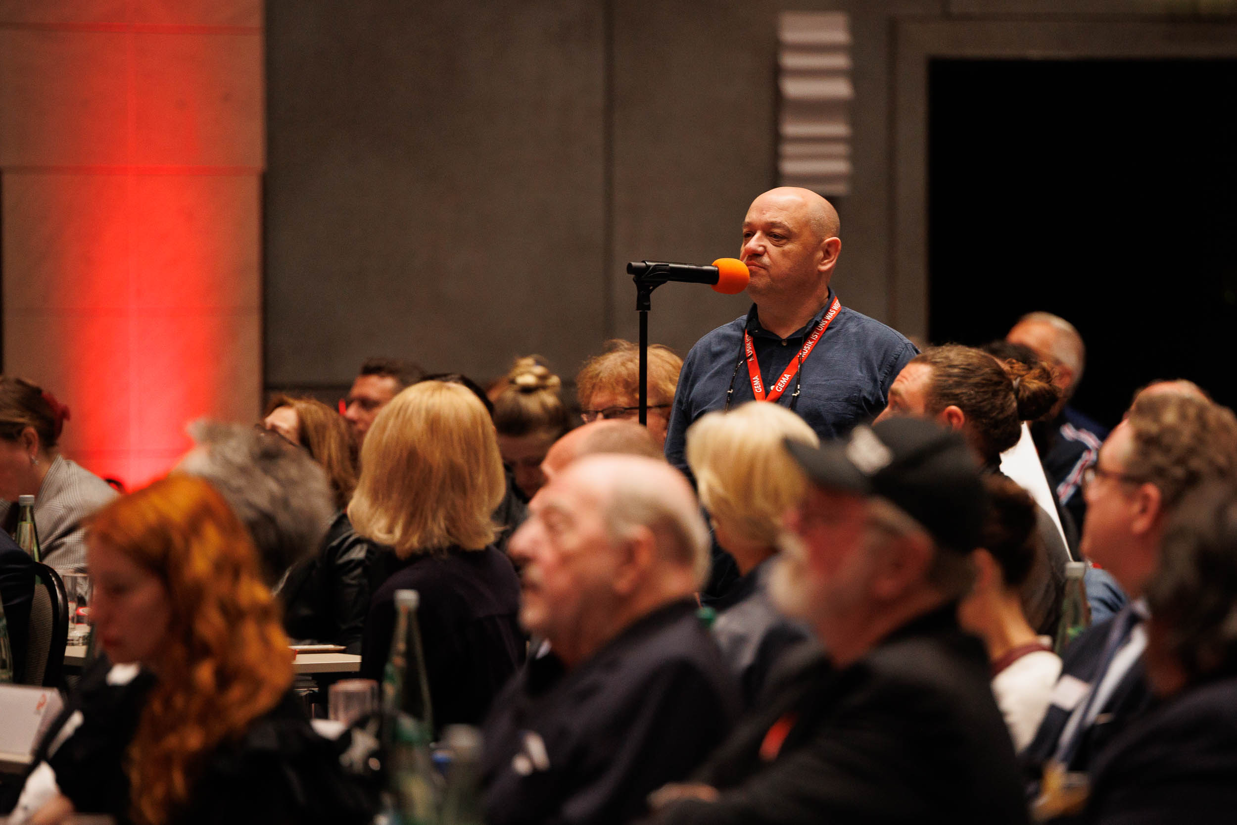 Ein Mann spricht in ein Mikrofon vor einem Publikum in einem Konferenzraum mit roter Beleuchtung.