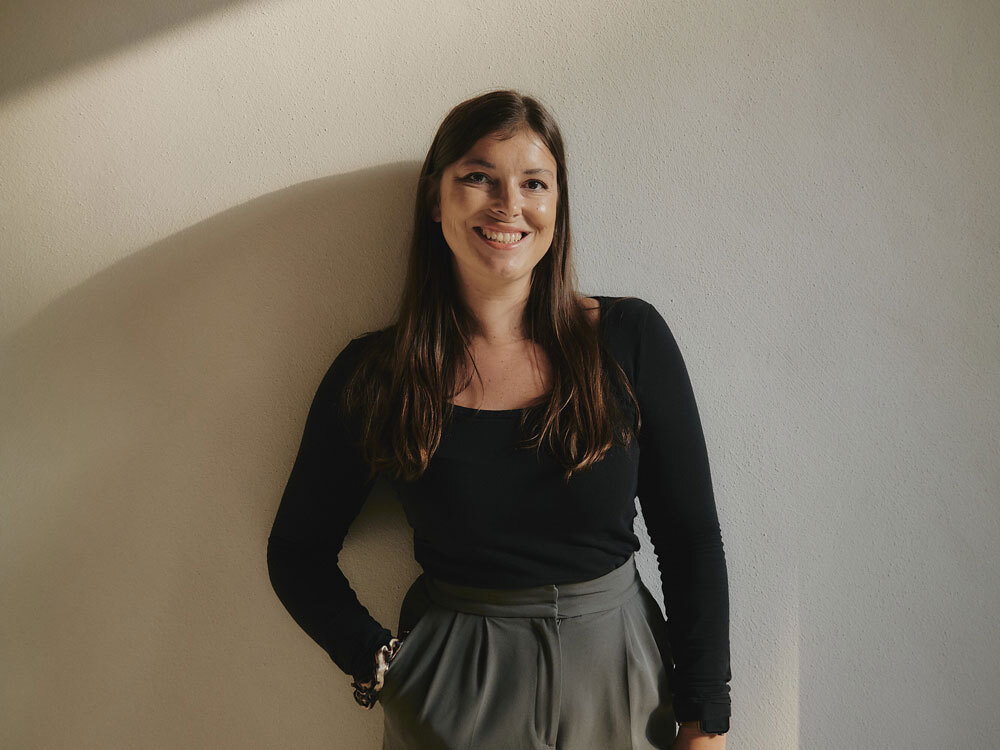 Eine lächelnde Frau in schwarzem Oberteil lehnt an einer hellen Wand.