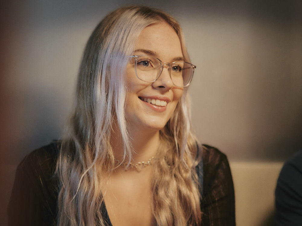 Eine lächelnde Person mit Brille und langen blonden Haaren in einem dunklen Oberteil.