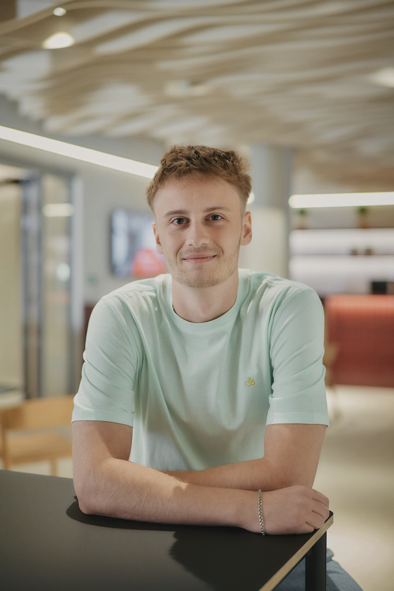 Ein Mann in einem mintgrünen T-Shirt sitzt lächelnd an einem Tisch in einem modernen Raum.