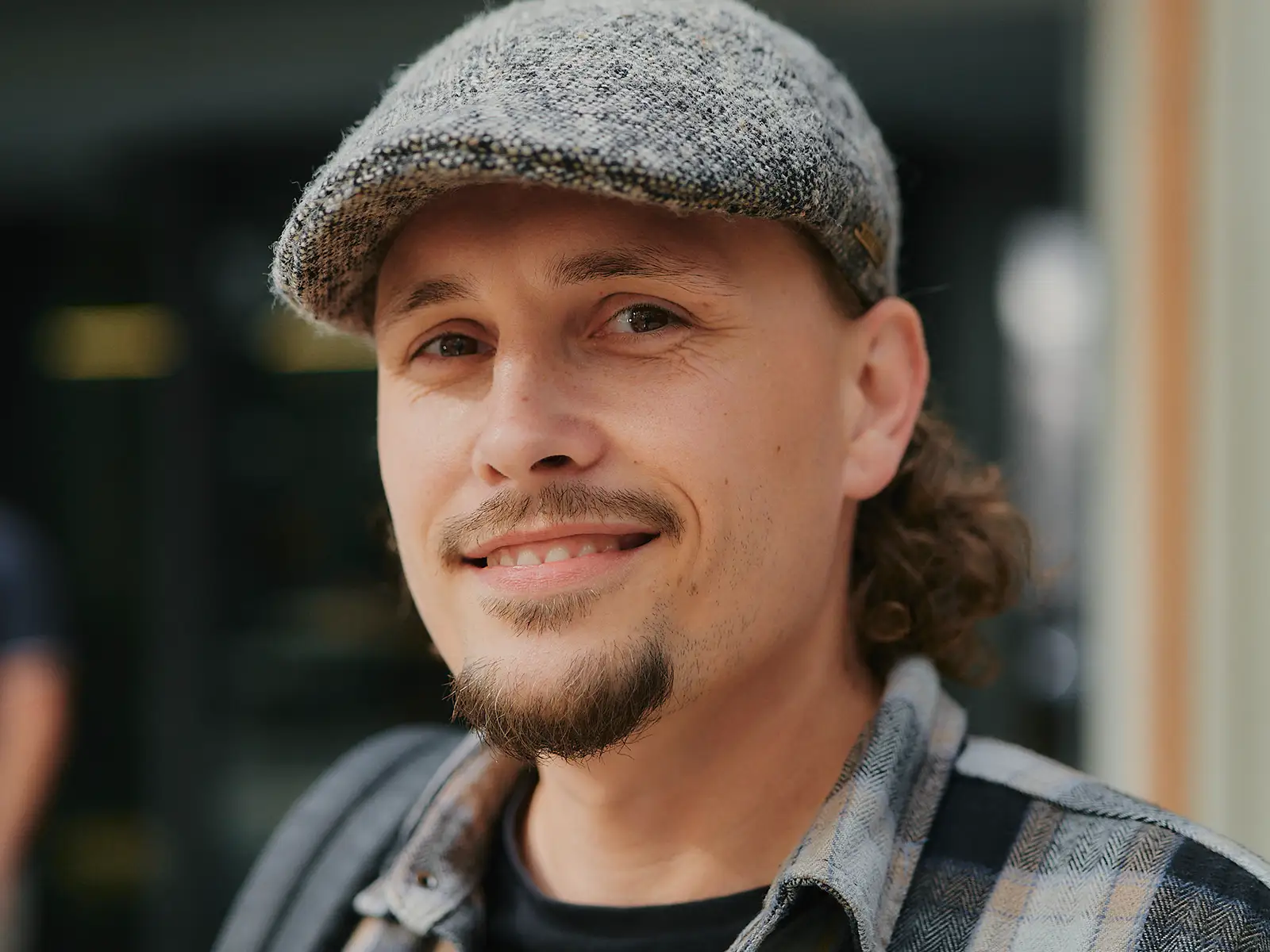 Ein lächelnder Mann mit Schnurrbart und grauer Schiebermütze trägt ein kariertes Hemd.