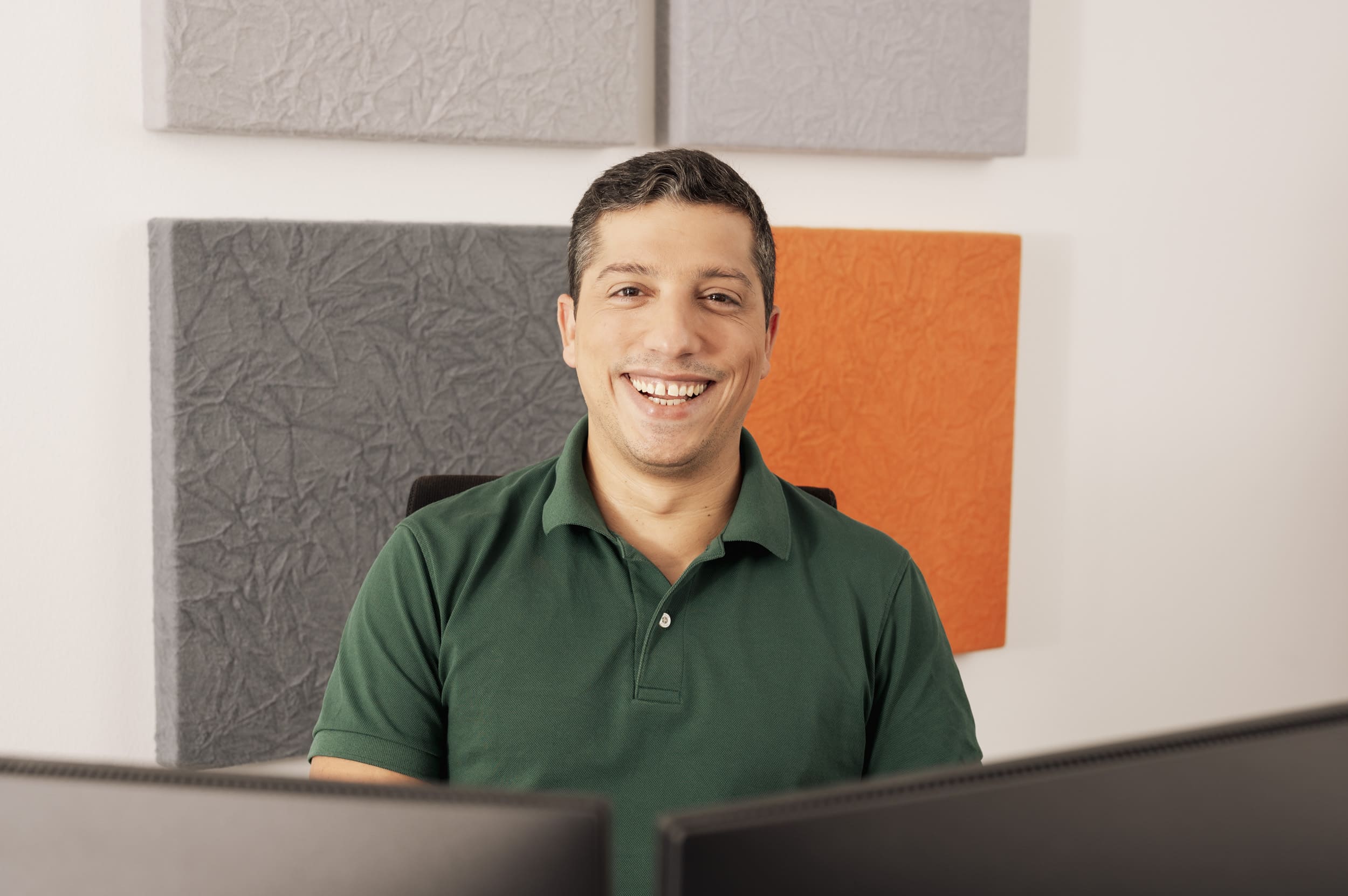 Ein lächelnder Mann in grünem Hemd sitzt vor grauen und orangefarbenen Akustikplatten.