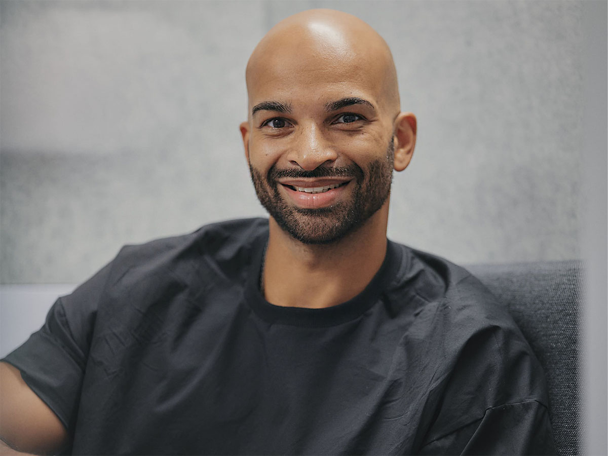 Ein lächelnder Mann mit Bart in einem schwarzen T-Shirt sitzt vor einem grauen Hintergrund.