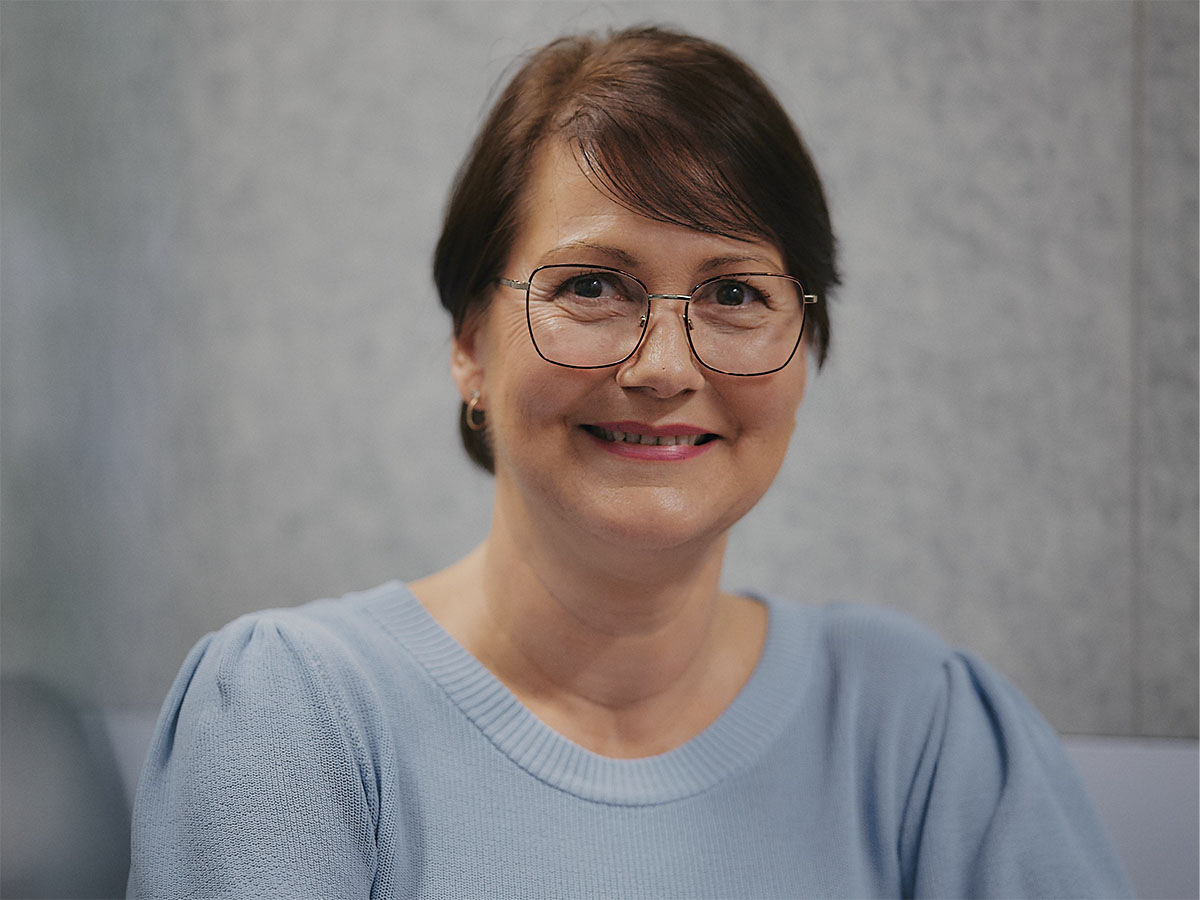 Eine Frau mit Brille und blauem Pullover lächelt vor einer grauen Wand.