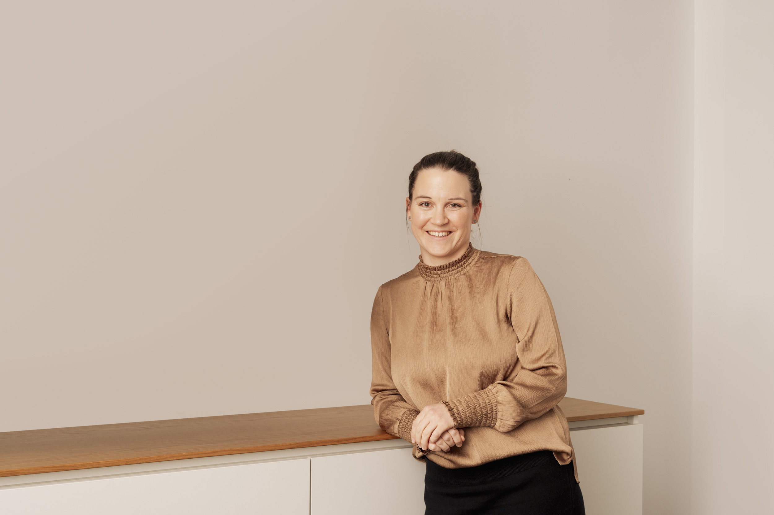 Eine lächelnde Frau in beiger Bluse lehnt an einem weißen Schrank vor beigem Hintergrund.
