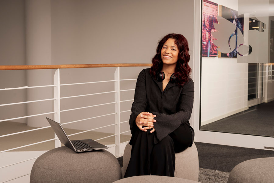 Eine lächelnde Person sitzt auf einem grauen Hocker neben einem Laptop in einem modernen Büro.