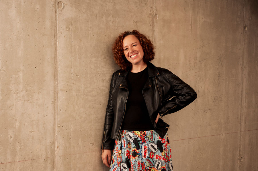 Eine lächelnde Person in Lederjacke und bunter Rock steht vor einer Betonwand.