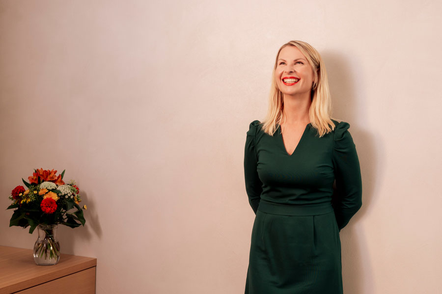 Frau in grünem Kleid lächelt vor beiger Wand, links ein Blumenstrauß auf einem Holztisch.