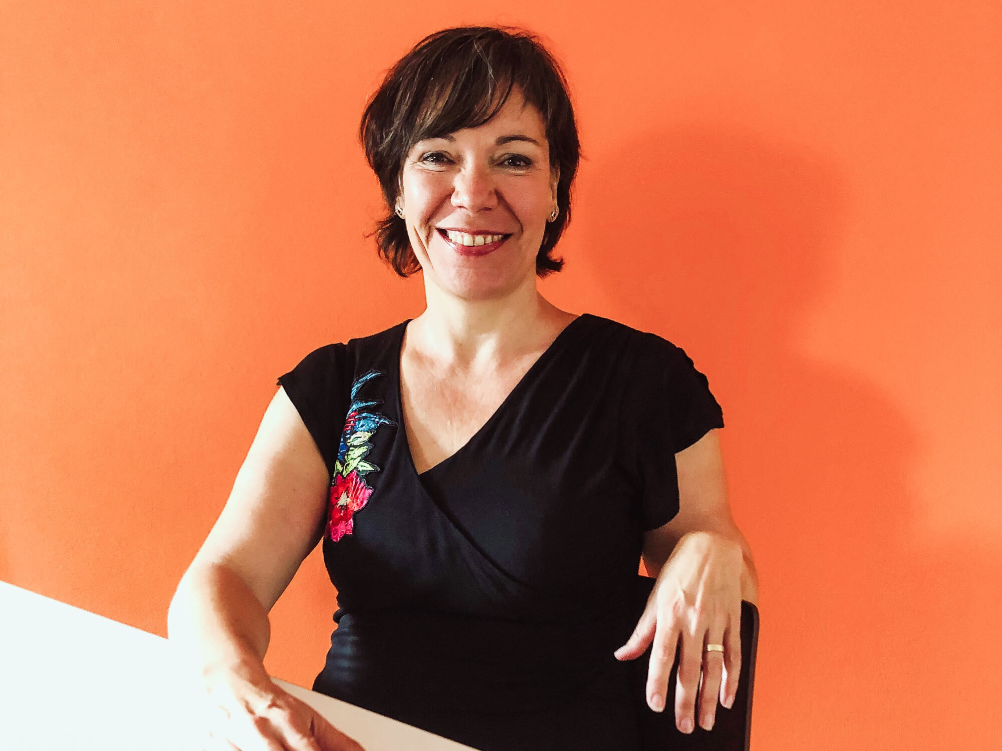 Eine lächelnde Frau in schwarzem Kleid sitzt vor einer orangefarbenen Wand.